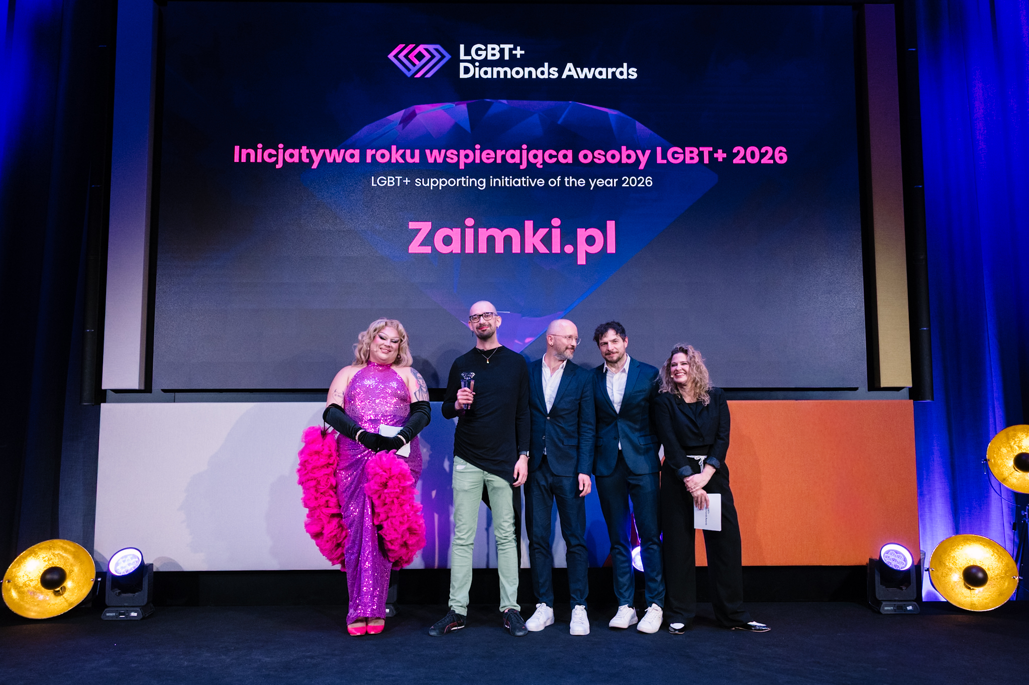 Na scenie stoją dwie osoby prezentujące, dwie wręczające nagrodę, oraz Andrea reprezentujące kolektyw; trzyma w dłoni statuetkę. W tle ekran z napisem "LGBT+ Diamonds Awards. Inicjatywa roku wspierająca osoby LGBT+ 2026. Zaimki.pl"